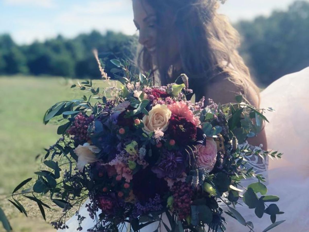 Braut im weißen Kleid hält einen üppigen Blumenstrauß in Rosa, Violett und Grün auf einer sonnigen Wiese.