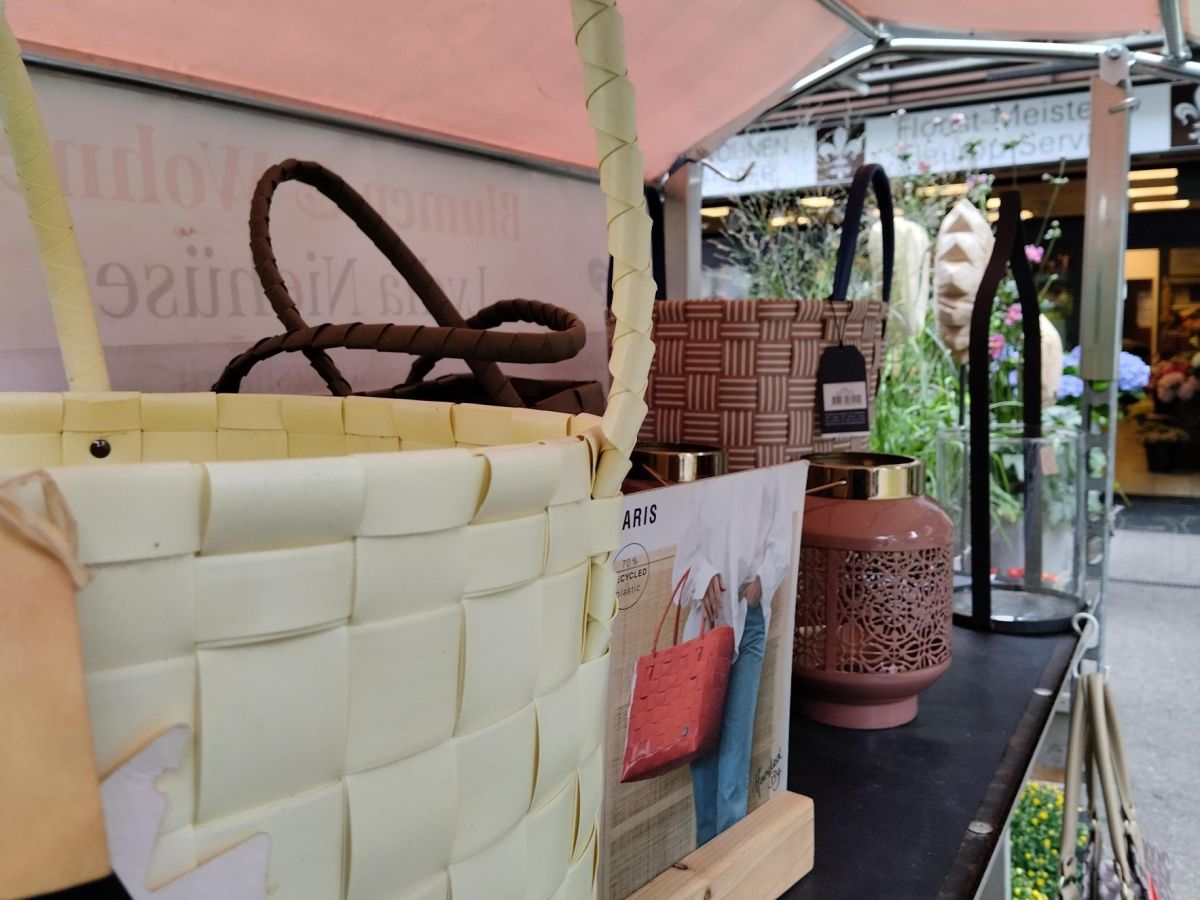 „Auslage mit geflochtenen Taschen, Laternen und Wohnaccessoires auf einem Tisch vor einem Blumenladen.