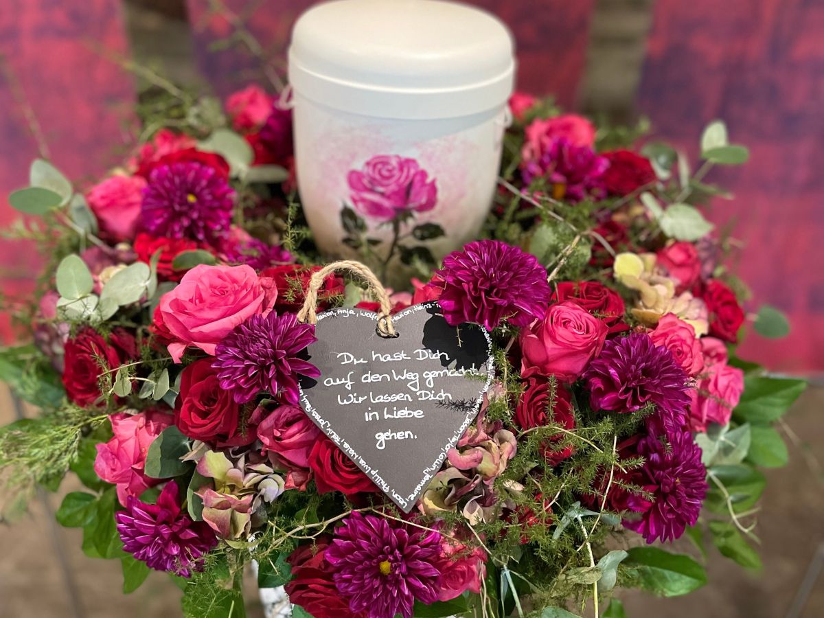 Runder Trauerkranz aus pinken und violetten Blumen mit Herzschild und Urne in der Mitte, liebevoll arrangiert.