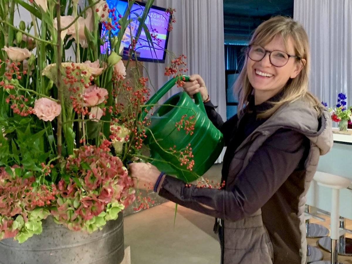 Floristin im Blumenladen hält einen bunten Blumenstrauß, im Hintergrund große dekorative Blüten und Wohnaccessoires.