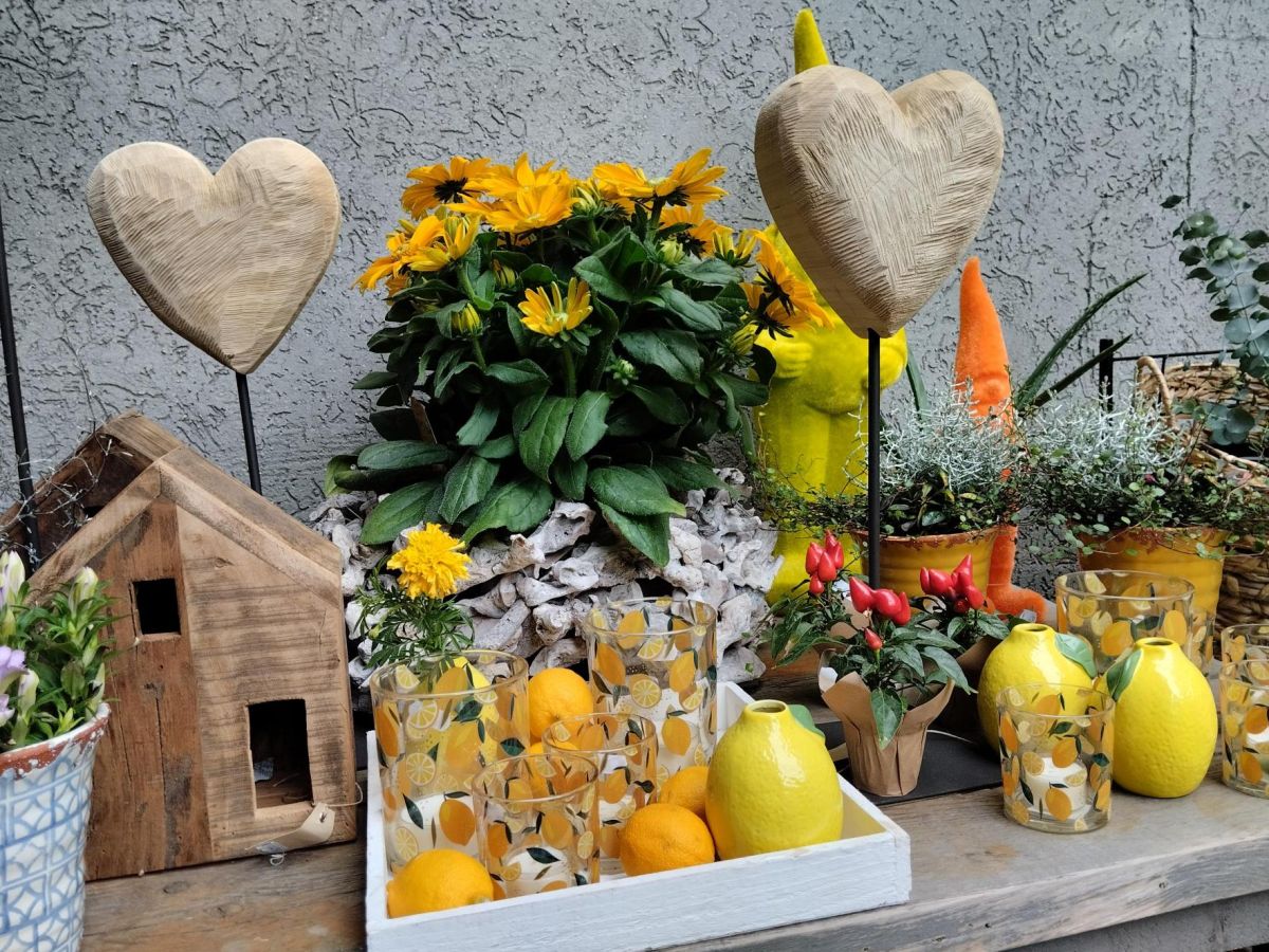 Dekoratives Arrangement mit gelben Blumen, Zitronen, Kerzenhaltern, Holzherzen und kleinen Pflanzen auf einem Regal.